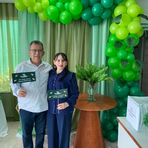 Um homem e uma mulher sorriem sob um arco de balões verdes, com plaquinhas da Essencial Pharma.