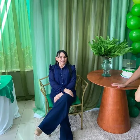 Uma mulher de azul-marinho sentada ao lado de uma mesa com uma planta, sob balões verdes.