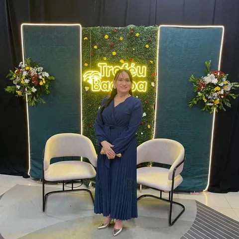 Uma mulher de vestido azul-marinho posa diante de um letreiro luminoso, entre poltronas e flores.